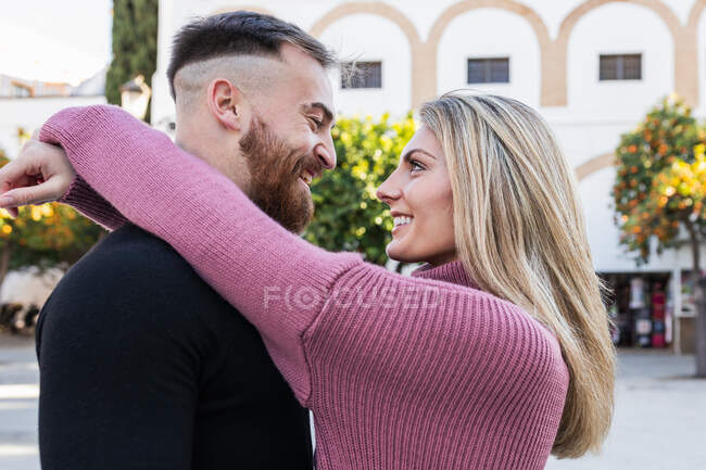 Vista lateral do homem terno abraçando a mulher encantadora sorridente enquanto estão juntos na cidade durante o passeio — Fotografia de Stock