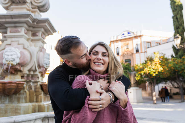 Zarter Mann küsst lächelnde charmante Frau auf die Wange, während er beim Stadtbummel zusammen steht — Stockfoto