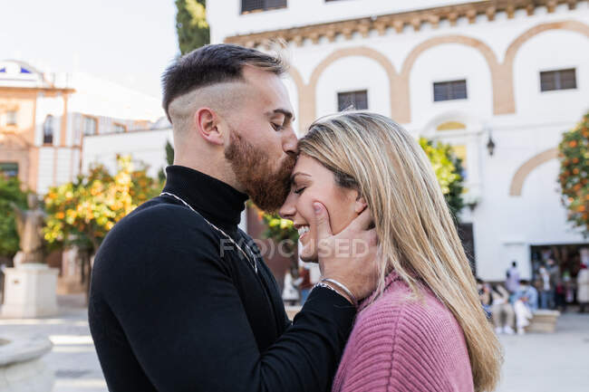 Side view of tender man kissing smiling charming woman in forehead while standing together in city during stroll — Stock Photo