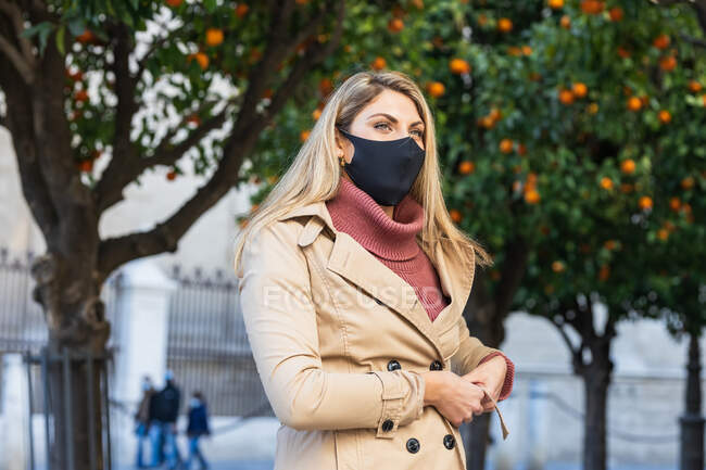 Tranquil female wearing protective mask and coat standing on city street during stroll and looking away — Stock Photo