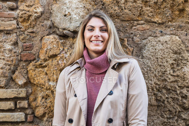 Zarte, charmante Frau in warmer Kleidung steht in der Stadtstraße auf einer steinernen alten Mauer und blickt in die Kamera — Stockfoto