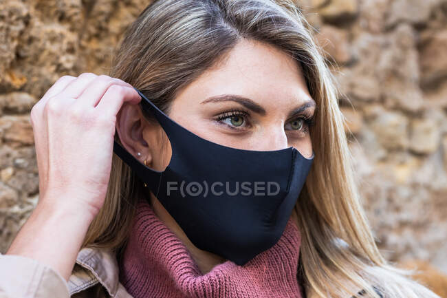 Kopfschuss einer zufriedenen Frau, die auf der Straße steht und eine Schutzmaske gegen Coronavirus aufsetzt, während sie wegschaut — Stockfoto