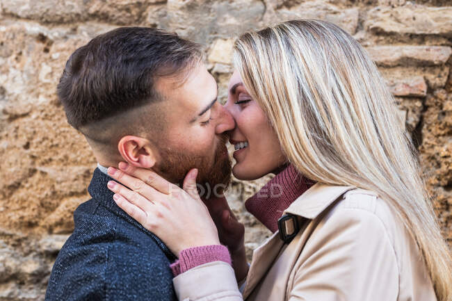 Side view of content couple gently kissing on city street against stone wall while enjoying weekend together — Stock Photo