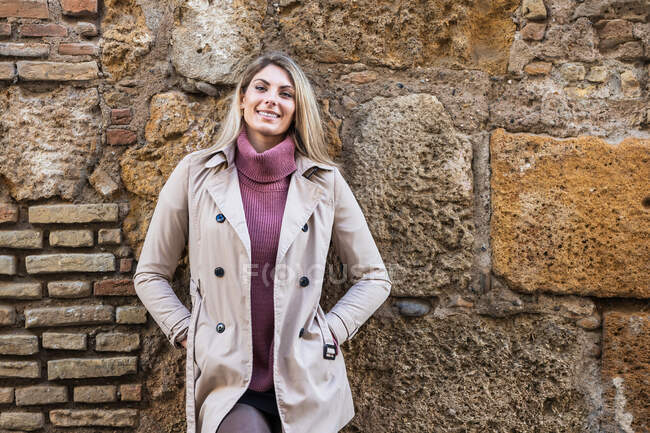 Tender charming female in warm clothes standing in city street on stone old wall looking at camera — Stock Photo