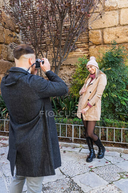 Novio con cámara fotográfica tomando foto de novia encantadora de pie en el parque urbano durante el paseo de fin de semana - foto de stock