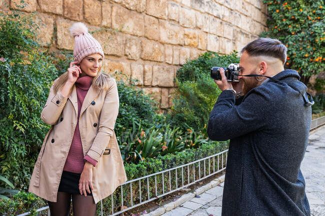 Парень с фотокамерой фотографирует очаровательную девушку, стоящую в городском парке во время прогулки по выходным — стоковое фото