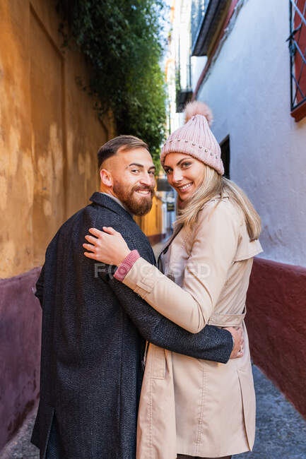 Vista trasera de la pareja enamorada en ropa de abrigo abrazando y caminando por la antigua calle estrecha mientras disfruta de un paseo por la ciudad y mirando a la cámara - foto de stock