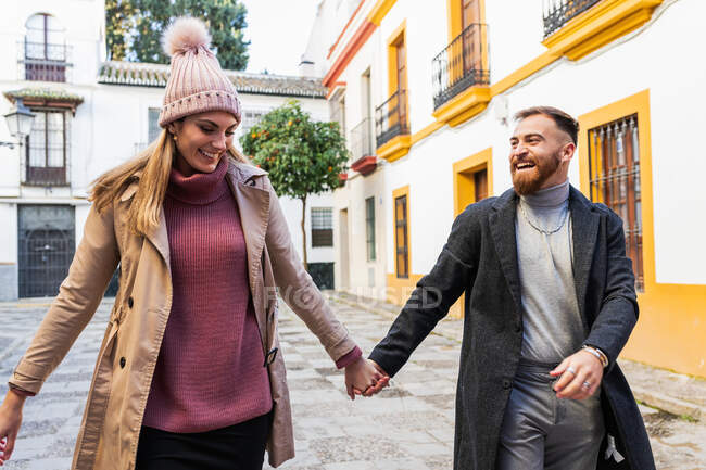 Задоволена пара в верхньому одязі тримає руки і ходить по вулиці, насолоджуючись вихідними разом у місті — стокове фото