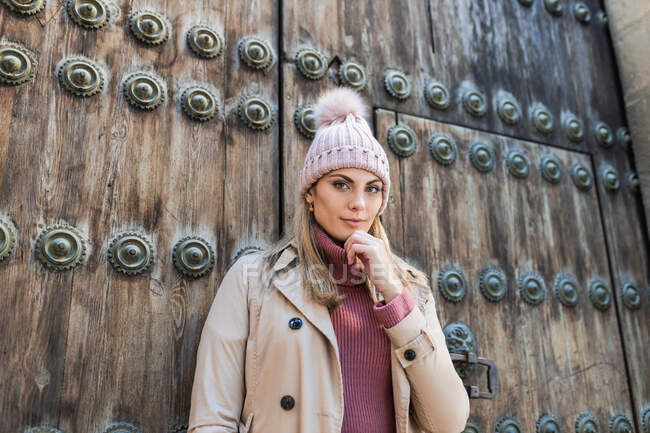 Низький кут вмісту жіночого пальто і капелюха, що стоїть біля старих дерев'яних дверей на вулиці і зворушливий підборіддя, дивлячись на камеру — стокове фото