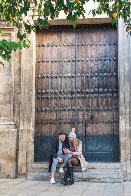 Couple aimant dans des vêtements de dessus élégants assis près d'énormes portes et se tenant la main tout en profitant week-end ensemble en ville — Photo de stock