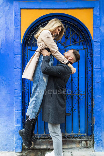 Вид збоку на зміст хлопець піднімає дівчину, стоячи на міській вулиці біля яскравої будівлі і дивлячись один на одного — стокове фото