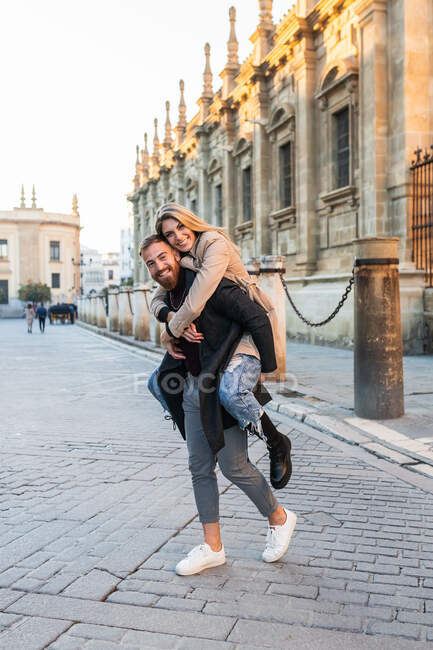 Juguetón hombre piggybacking mujer alegre mientras se divierten en la calle de la ciudad y mirando a la cámara - foto de stock