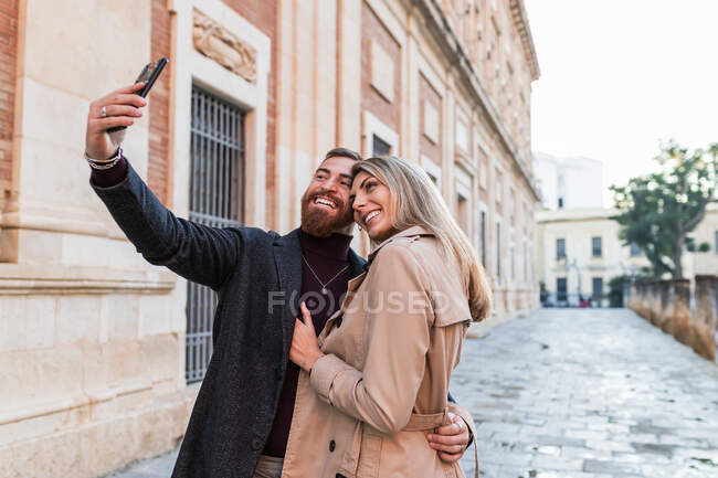 Любящая супружеская пара в модной верхней одежде стоит на улице и делает самоснимок на смартфоне во время прогулки по выходным — стоковое фото