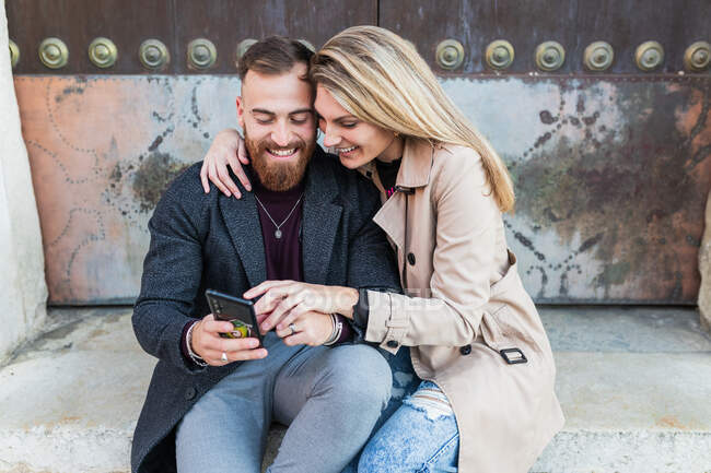 Couple aimant dans des vêtements de dessus élégants assis près de vieilles portes naviguant sur le téléphone mobile tout en profitant week-end ensemble en ville — Photo de stock