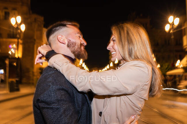 Vista lateral de pareja despreocupada abrazándose mirándose en la ciudad por la noche - foto de stock