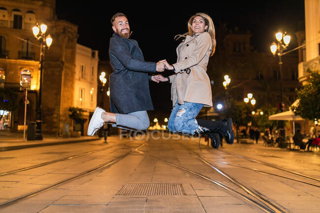 Vista lateral do casal despreocupado de mãos dadas e pulando acima da estrada enquanto se diverte na cidade à noite e olhando para a câmera — Fotografia de Stock