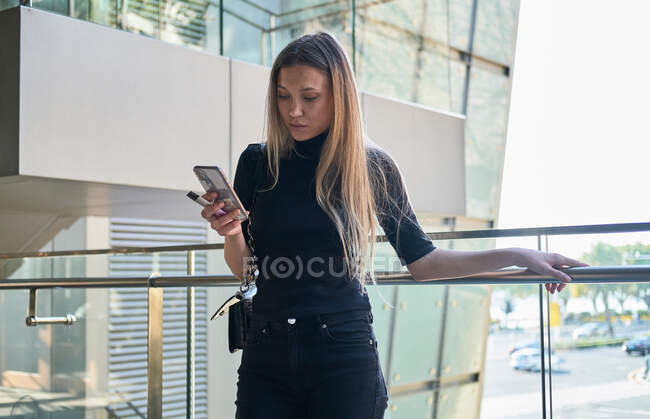 Cultivo de hermosa chica joven rubia usando su teléfono inteligente mirando preocupado con ropa negra - foto de stock