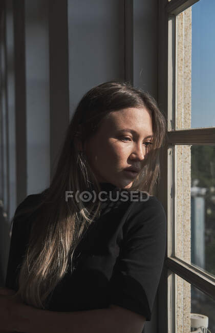 Retrato de menina bonita com cabelo loiro longo, ela está perto da janela e a luz do sol está iluminando seu rosto. — Fotografia de Stock