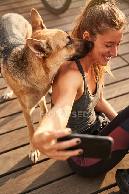 Сверху улыбающаяся женщина, сидящая рядом с немецкой овчаркой во время перерыва в беговой тренировке и фотографирующая себя на мобильном телефоне — стоковое фото