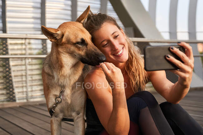 Mujer sonriente sentada cerca del perro pastor alemán durante el descanso en el entrenamiento para correr y tomar autorretrato en el teléfono móvil - foto de stock