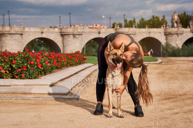 Cuerpo completo de corredora positiva besando perro pastor alemán durante el entrenamiento activo en el parque - foto de stock
