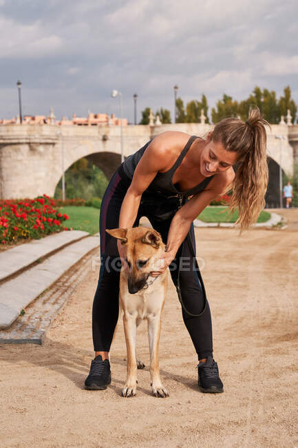 Corpo inteiro de corredor feminino positivo acariciando cão pastor alemão durante o treinamento ativo no parque — Fotografia de Stock