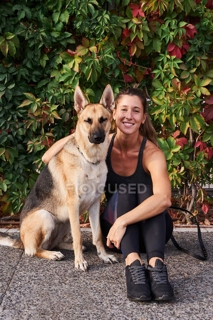 Corpo inteiro de corredor feminino positivo abraçando cão pastor alemão durante o treinamento ativo no parque — Fotografia de Stock