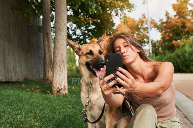 Mujer enfocada con el pelo largo sentado en la hierba cerca de pastor alemán mascota y tomar una selfie - foto de stock