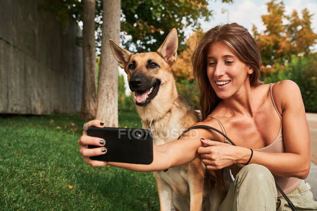 Konzentrierte Frau mit langen Haaren sitzt im Gras neben Schäferhund-Haustier und macht ein Selfie — Stockfoto