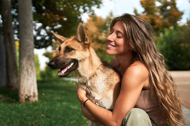 Propietaria femenina positiva abrazando perro pastor alemán mientras están sentados juntos en el césped en el parque - foto de stock