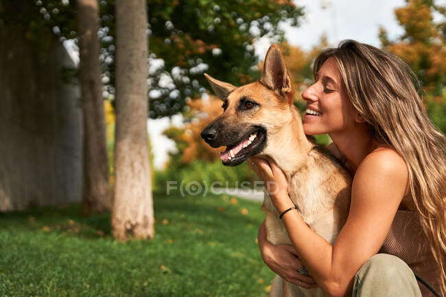 Propietaria femenina positiva abrazando perro pastor alemán mientras están sentados juntos en el césped en el parque - foto de stock