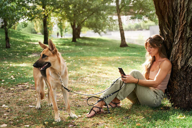 Женщина с длинными волосами сидит на траве рядом с немецкой овчаркой и пользуется мобильным телефоном в парке — стоковое фото