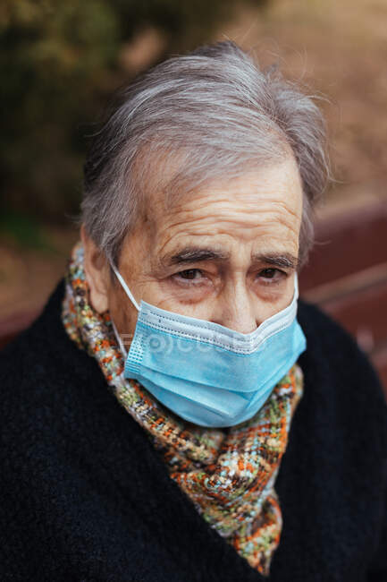 Porträt einer älteren Frau mit Gesichtsmaske, die auf einer Parkbank in die Kamera blickt — Stockfoto