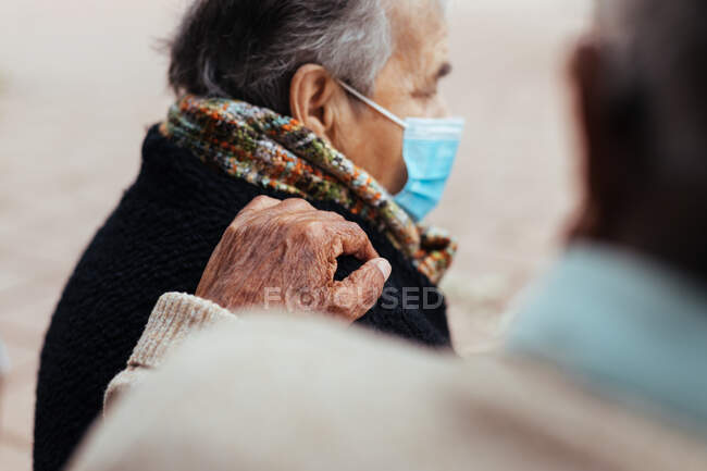Seitenansicht eines älteren Ehepaares, das auf einer Parkbank sitzt, während der Ehemann seine Hand in einer Geste der Liebe auf die Schulter seiner Frau legt — Stockfoto