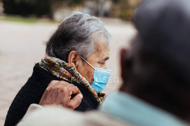 Vista lateral de una pareja de ancianos sentados en un banco del parque mientras el marido pone su mano sobre el hombro de su esposa en un gesto de amor - foto de stock