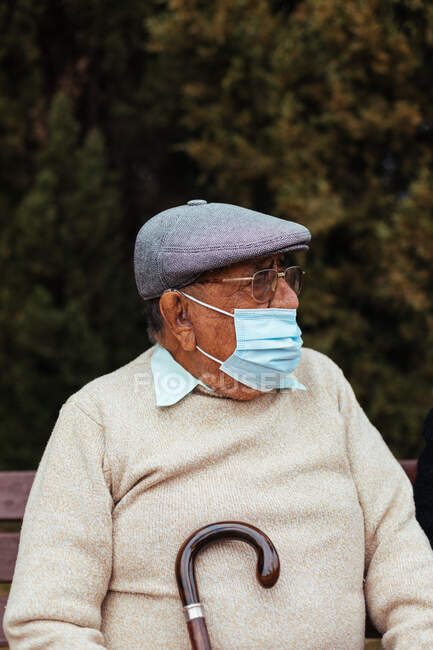 Ritratto di uomo anziano con maschera facciale e basco che distoglie lo sguardo seduto su una panchina del parco — Foto stock