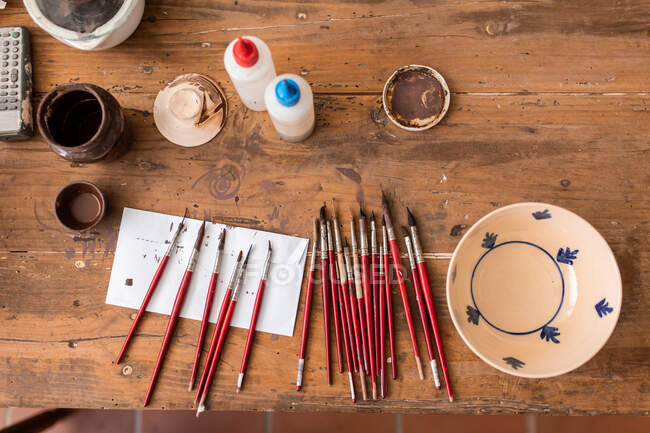 From above of various brushes near painted ceramic plate on wooden table in workshop — Stock Photo