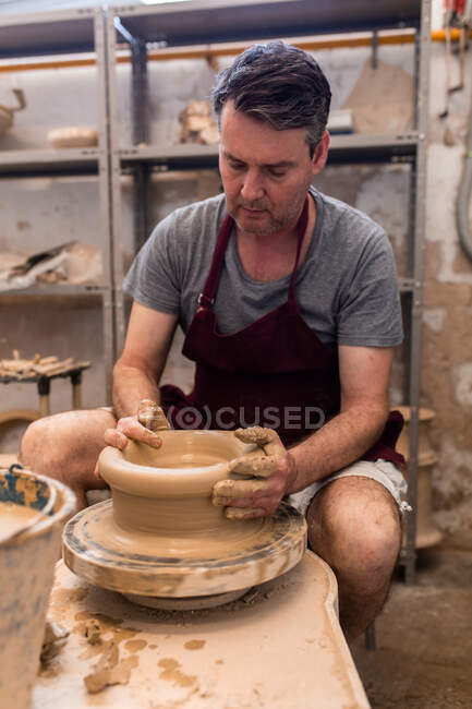 Corpo pieno di maestro maschio concentrato in grembiule seduto a tavola mentre scolpisce con argilla marrone sulla ruota di lancio — Foto stock