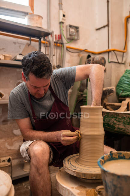 Full body of concentrated male master in apron sitting at table while sculpting with brown clay on throwing wheel — Stock Photo