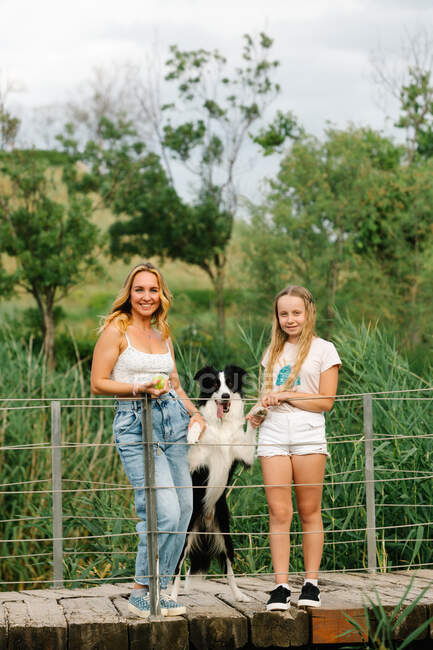 Content mother and teenage daughter standing with Border Collie dog on wooden footbridge in summer park and looking at camera — Stock Photo