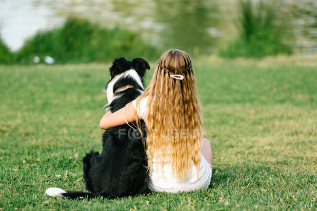 Обратный вид на неузнаваемую девочку-подростка, обнимающую пушистую собаку Пограничного колли, сидящую на лужайке возле пруда в летнем парке — стоковое фото
