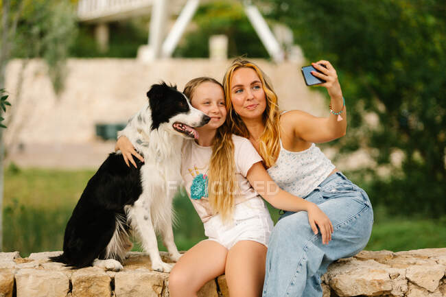 Радісна мати з донькою сидять на кам'яному паркані разом з Френдлі Борд Коллі собакою і б'ються на смартфоні в літній парк — стокове фото