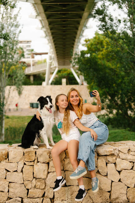 Мать и дочь сидят на каменном заборе с собакой бордер-колли и делают селфи на смартфон в летнем парке — стоковое фото