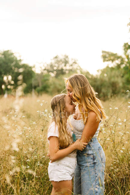 Vue latérale de la mère aimante embrassant tendrement adolescente dans le front tout en câlinant et debout dans la prairie en été — Photo de stock