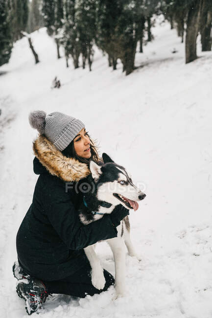 Молода етнічна леді, вдягнена в сукню, обіймає гарненького лагідного пса, коли взимку сидить у сніжних лісах біля зелених сосків. — стокове фото