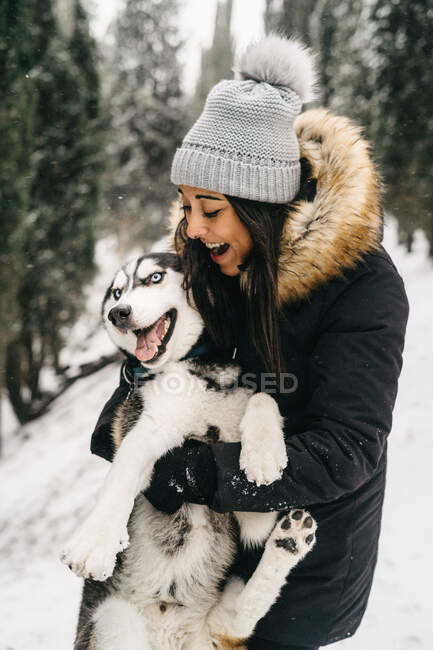 Smiling young ethnic lady wearing outerwear hugging cute husky dog while standing in snowy woods near green spruces in winter day — Stock Photo