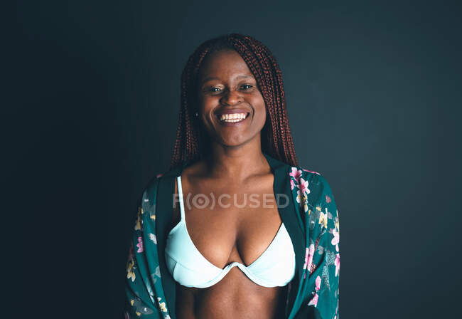 Delighted African American female in bra and dressing gown standing on black background in studio and looking at camera — Stock Photo
