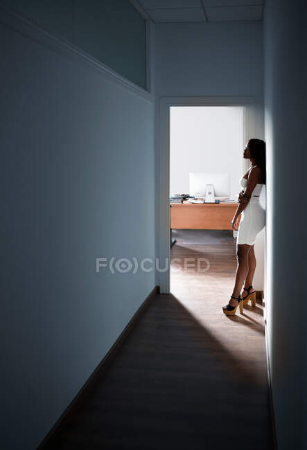Side view of female in dress and high heels leaning on wall while standing in corridor in modern apartment — Stock Photo