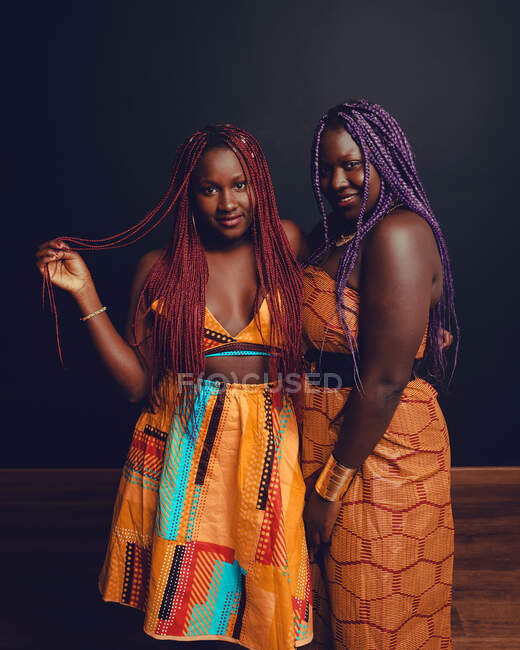 Zufriedene afroamerikanische Freundinnen mit bunten Zöpfen und in leuchtend orangefarbener Kleidung stehen vor dunklem Hintergrund im Studio und schauen in die Kamera — Stockfoto