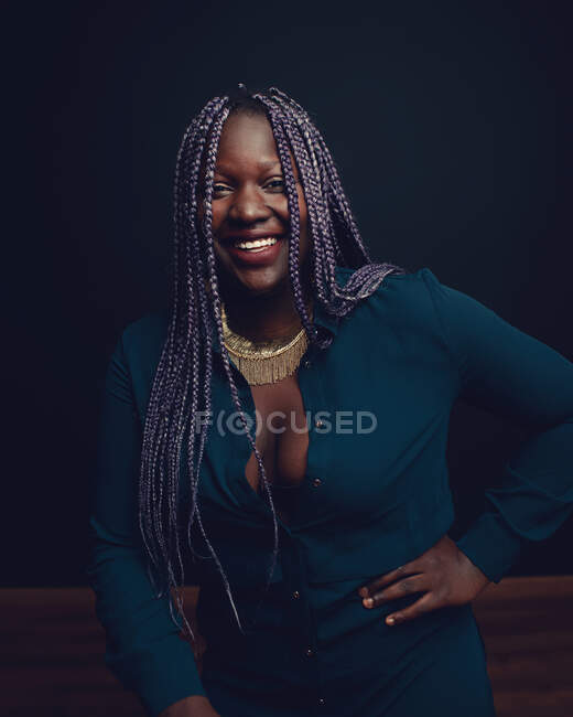 Cheerful African American female with black braids standing on black background in studio and looking at camera — Stock Photo